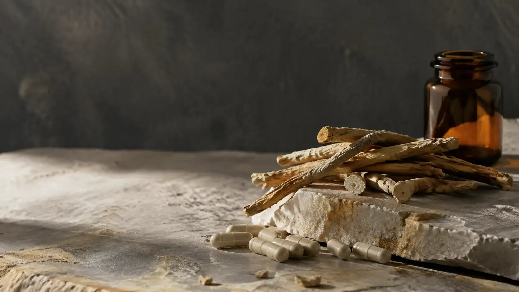 Dried ashwagandha root, whole and cut, resting on pale mineral stone beside unbranded capsules in cold daylight.